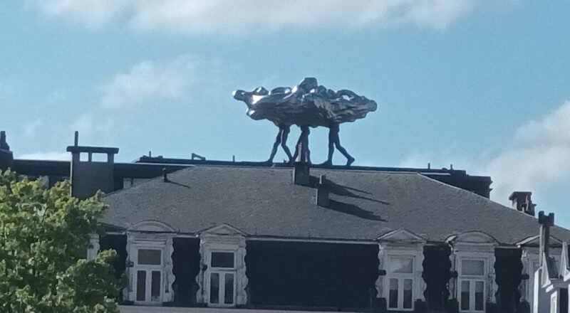 Escultura metálica de figuras humanas cargando una forma abstracta brillante, ubicada sobre el techo de un edificio clásico bajo un cielo azul con nubes.