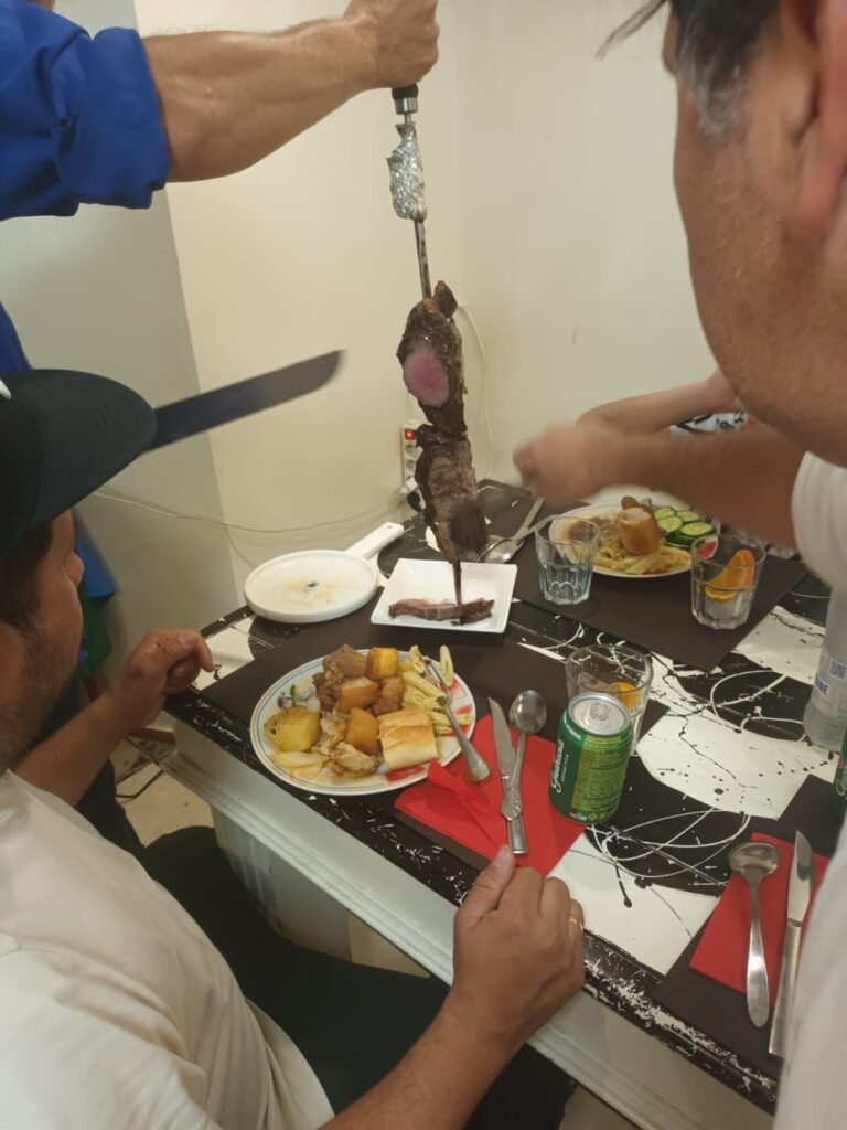 Un camarero sirve carne asada directamente a los clientes en una mesa decorada con detalles brasileños, como panes y ensaladas.